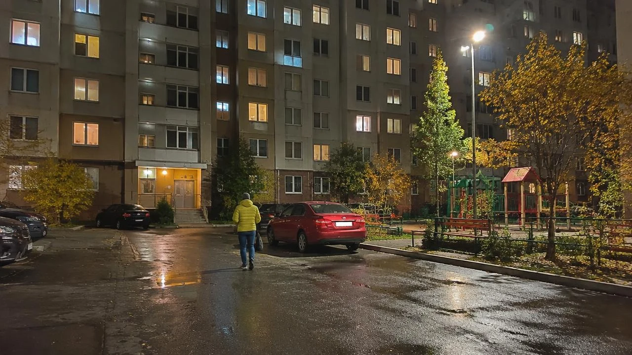 Ночной двор панельного дома в цвете: красная машина и человек в жёлтой куртке под фонарём